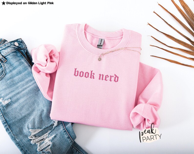 Op de afbeelding: Een lichtroze sweatshirt met de geborduurde tekst "book nerd" in een gotisch lettertype. Het sweatshirt wordt getoond met een spijkerbroek met distress details en een delicate ketting. De woorden "peak party" staan rechtsonder.