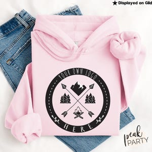 May include: A light pink hoodie with a black circular graphic featuring crossed arrows, trees, a mountain, and a campfire. The text "YOUR OWN LOGO HERE" is printed around the graphic. The hoodie is displayed on blue jeans.