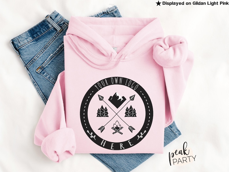 May include: A light pink hoodie with a black circular graphic featuring crossed arrows, trees, a mountain, and a campfire. The text "YOUR OWN LOGO HERE" is printed around the graphic. The hoodie is displayed on blue jeans.