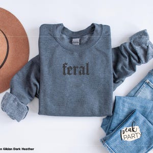 May include: A dark heather gray sweatshirt with the word "feral" embroidered in a gothic font. The sweatshirt is laid flat with a brown hat and blue jeans. The text "peak PARTY" is on the jeans.