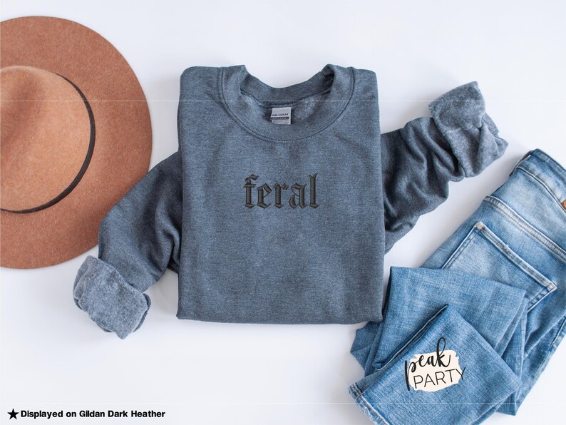May include: A dark heather gray sweatshirt with the word "feral" embroidered in a gothic font. The sweatshirt is laid flat with a brown hat and blue jeans. The text "peak PARTY" is on the jeans.