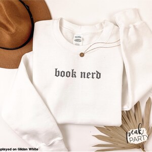 Op de afbeelding: Een witte crewneck sweatshirt met de tekst "book nerd" in een gotisch lettertype. Een gouden ketting en een bruine hoed liggen naast de sweatshirt. De sweatshirt wordt getoond op Gildan White.
