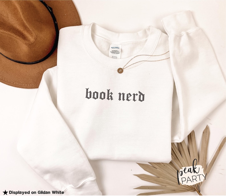 Op de afbeelding: Een witte crewneck sweatshirt met de tekst "book nerd" in een gotisch lettertype. Een gouden ketting en een bruine hoed liggen naast de sweatshirt. De sweatshirt wordt getoond op Gildan White.