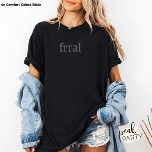 May include: Black t-shirt with the word "feral" in a gothic font. The shirt is displayed on a person wearing a denim jacket and shorts. The text "Displayed on Comfort Colors Black" is at the top left.