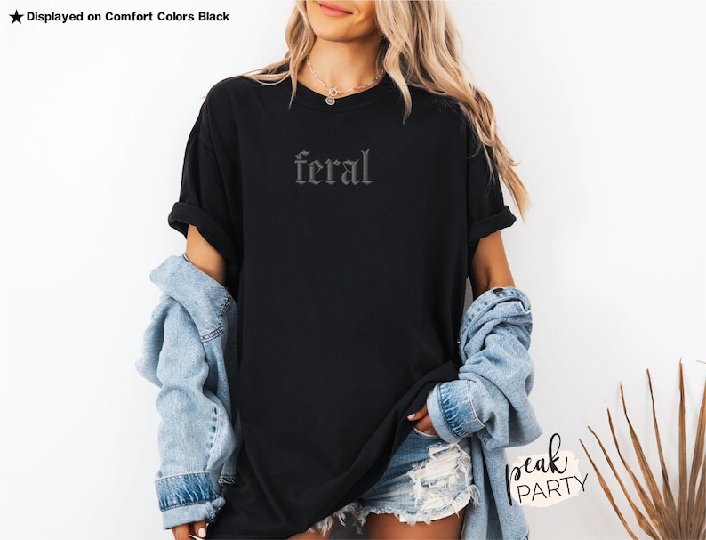 May include: Black t-shirt with the word "feral" in a gothic font. The shirt is displayed on a person wearing a denim jacket and shorts. The text "Displayed on Comfort Colors Black" is at the top left.