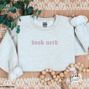 Op de afbeelding: Een lichtgrijs sweatshirt met de geborduurde tekst "book nerd" in een lichtpaarse gotische letter. Het sweatshirt wordt gepresenteerd op een getextureerd oppervlak met decoratieve elementen, waaronder groen en houten kralen.