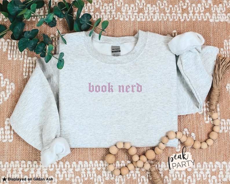 Op de afbeelding: Een lichtgrijs sweatshirt met de geborduurde tekst "book nerd" in een lichtpaarse gotische letter. Het sweatshirt wordt gepresenteerd op een getextureerd oppervlak met decoratieve elementen, waaronder groen en houten kralen.