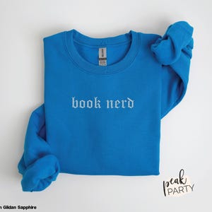 Op de afbeelding: Een blauwe sweatshirt met de woorden "book nerd" in witte gotische letters. De sweatshirt is opgevouwen, met de mouwen opgerold. Het merklabel is zichtbaar. De woorden "Peak Party" staan rechtsonder.
