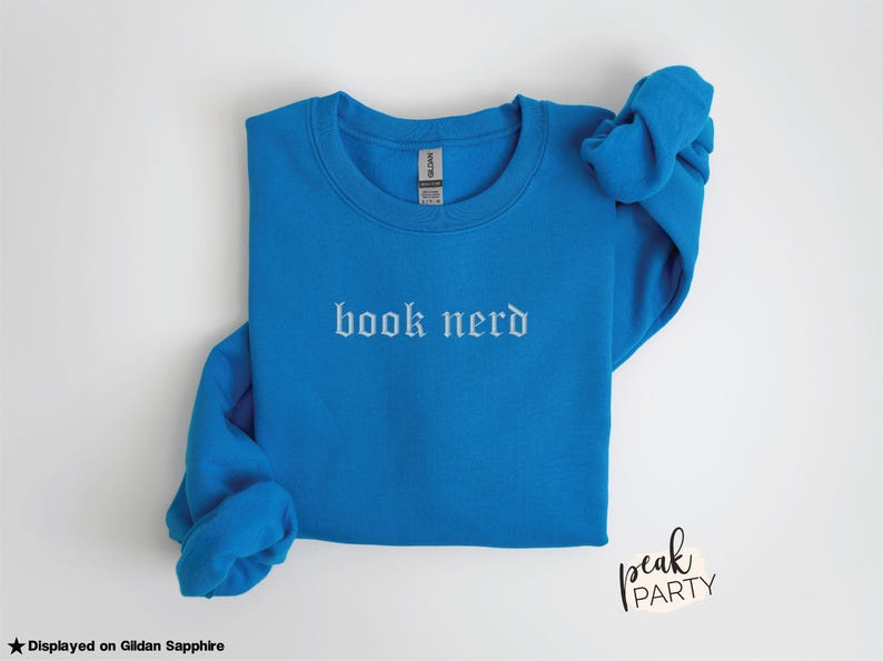 Op de afbeelding: Een blauwe sweatshirt met de woorden "book nerd" in witte gotische letters. De sweatshirt is opgevouwen, met de mouwen opgerold. Het merklabel is zichtbaar. De woorden "Peak Party" staan rechtsonder.