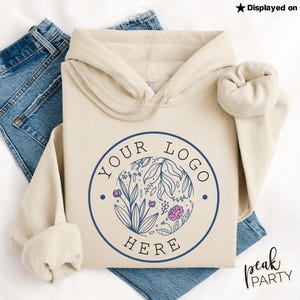 May include: Cream-colored hoodie with a circular logo featuring floral and leaf designs in blue and pink, with the text "YOUR LOGO HERE". The hoodie is displayed on top of blue jeans. The text "Displayed on Gildan Sand" is visible.