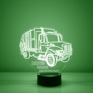 May include: A white silhouette of a garbage truck with the text "DAVIS WASTE MANAGEMENT" below it. The truck is illuminated from behind, creating a glowing effect. The truck is on a black base.