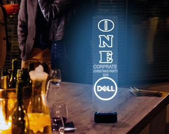 May include: A clear acrylic sign with the word "ONE" in white letters, followed by "CORPORATE CHRISTMAS PARTY 2020" and the Dell logo. The sign is lit from behind, creating a glowing effect.