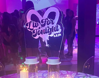 May include: A table setting with a white tablecloth featuring a colorful heart pattern. There are several clear glass cylinders with white candles and a rainbow number "5" on the table. A clear acrylic sign with the text "Liv for Tonight 6.8.24" is in the background.