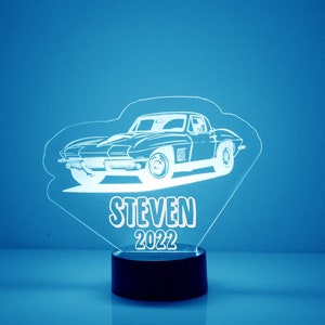 May include: A white silhouette of a classic car with the name "STEVEN" and the year "2022" below it. The car is on a clear acrylic base with a black round base.