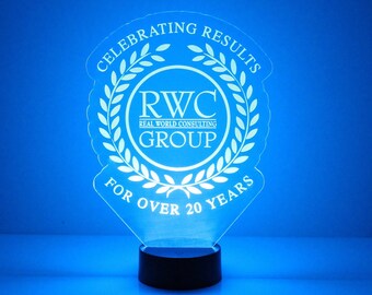 May include: A clear acrylic award with a white wreath design and the text "Celebrating Results" and "For Over 20 Years". The center of the award features a white circle with the text "RWC Real World Consulting Group". The award is on a black base.