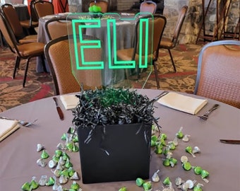 May include: A green acrylic sign with the name "ELI" on it, sitting on a black box filled with black shredded paper. The box is surrounded by silver and green foil-wrapped chocolate kisses on a gray tablecloth.
