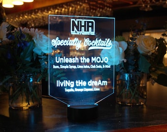May include: A blue illuminated sign with white text that reads "NHA Twelve & Twenty Two" "Specialty Cocktails" "Unleash the MOJO Rum, Simple Syrup, Lime Juice, Club Soda, & Mint" "living the dream Tequila, Orange Liqueur, Lime"