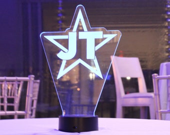May include: A clear acrylic star-shaped centerpiece with the initials "JT" in white lettering. The base is black and the star is illuminated with a white light.