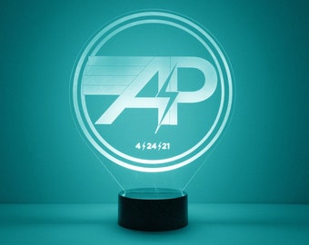 May include: A white acrylic night light with a circular base and a clear acrylic shade. The shade features a stylized graphic of the letters "AP" with a lightning bolt in the middle. The date "4/24/21" is printed below the graphic.