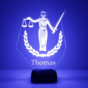 May include: A white acrylic plaque with a silhouette of the figure of justice holding scales and a sword. The plaque has a laurel wreath design and the name "Thomas" is printed below the figure.