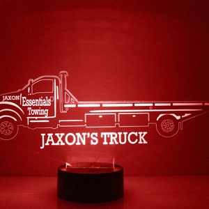 Könnte beinhalten: Weiße Silhouette eines Abschleppwagens mit dem Text "Jaxon's Essentials Towing" und "Jaxon's Truck" auf rotem Hintergrund. Der Wagen steht auf einem schwarzen Sockel.