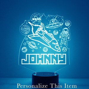 May include: A white space-themed night light featuring a rocket ship, planets, stars, an astronaut, and a UFO. The name "JOHNNY" is written in block letters at the bottom of the light.
