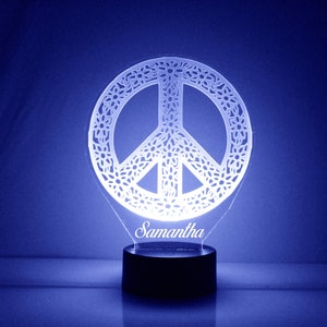 Puede incluir: Un signo de paz blanco con un diseño floral, iluminado con una luz blanca. El nombre Samantha está escrito debajo del signo de la paz.