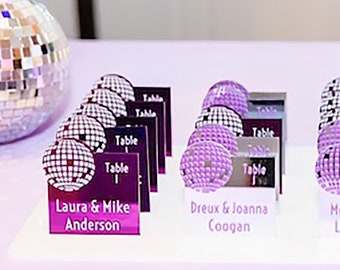 May include: Purple and silver table number cards with a disco ball design. The cards have the text "Table 1" and the names "Laura & Mike Anderson", "Dreux & Joanna Coogan", and "M L".