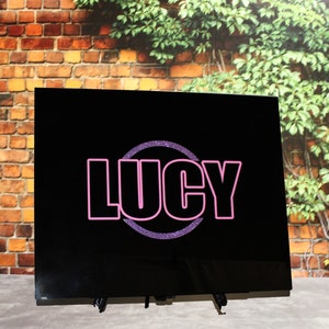 May include: Black sign with a pink glitter circle and the name "LUCY" in pink outlined letters.