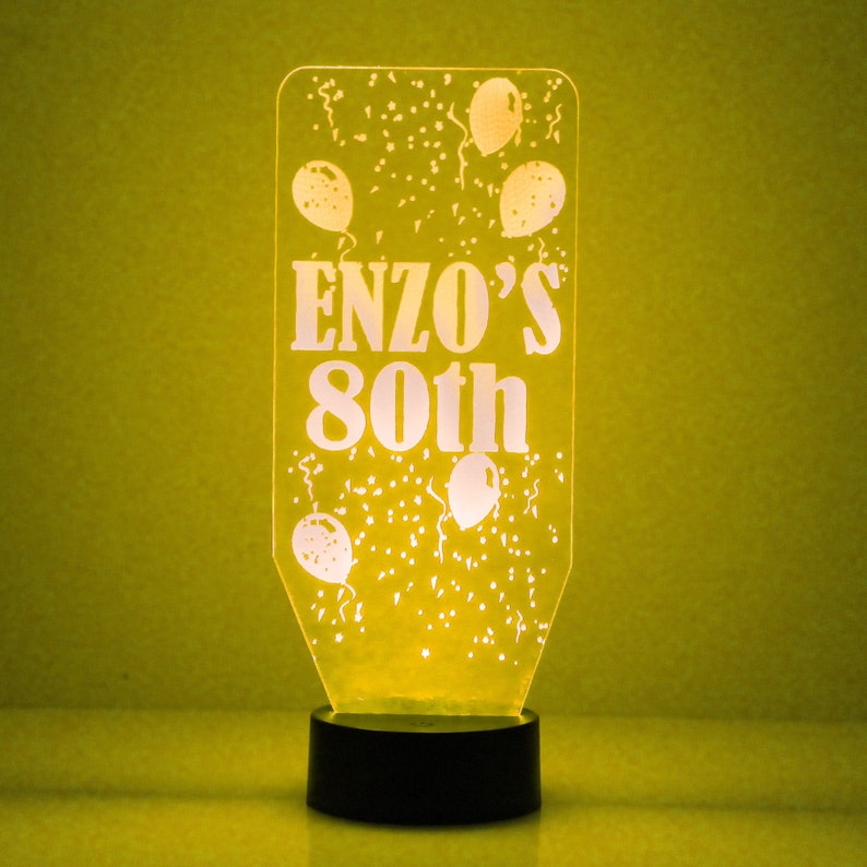 May include: A clear acrylic night light with a white base. The light features a design of balloons and confetti with the text "ENZO'S 80th".