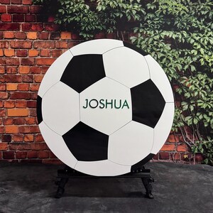 May include: A white and black soccer ball with the name "JOSHUA" printed in green on the front. The ball is on a black stand.