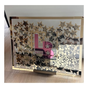 May include: A gold-framed mirror with a pink letter "B" in the center, surrounded by gold star-shaped cutouts. The stars have handwritten messages. The date "10.11.25" is visible. The mirror is displayed on a wooden surface.