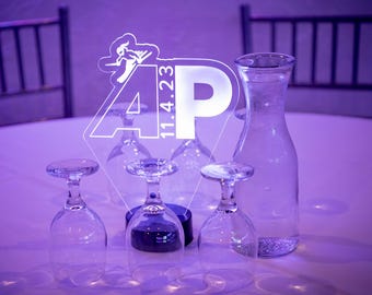 May include: A clear acrylic sign with the letters "AP" and the date "11.4.23" in white. The sign is illuminated from behind, casting a purple glow. The sign is on a table with four clear glass goblets and a clear glass pitcher.
