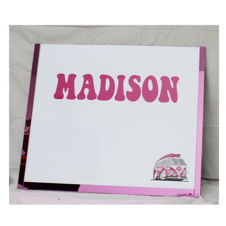 May include: White dry erase board with a pink frame. The board has the name "MADISON" written in pink letters and a pink and white cartoon van with a surfboard on top.
