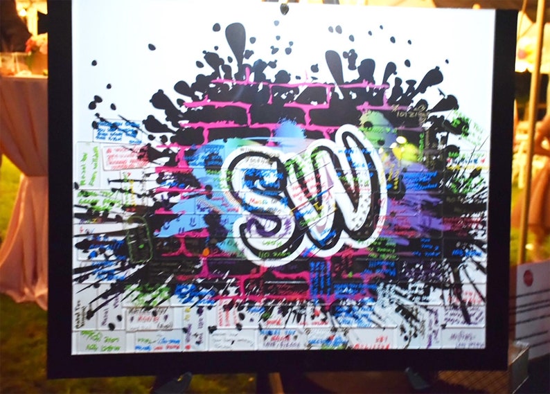 May include: A white canvas with a black brick wall design and a large graffiti-style "SW" in white. The canvas is covered in colorful paint splatters and has a black frame.