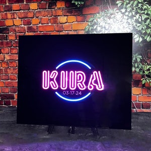 Op de afbeelding: Een zwart bord met een roze neon-bord met de tekst "KURA" in een cirkel met een blauwe neon-omlijsting. Het bord heeft ook de tekst "03:17:24" onder de cirkel.