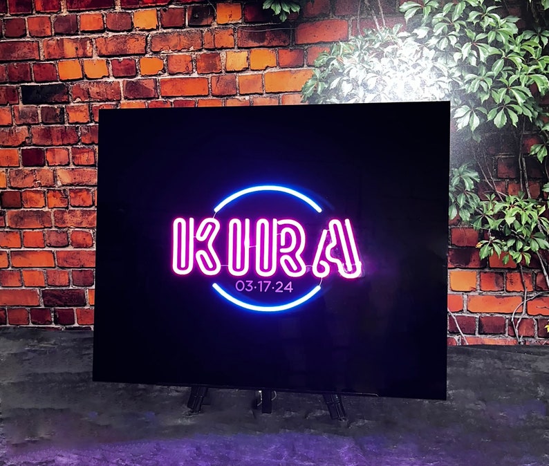 Op de afbeelding: Een zwart bord met een roze neon-bord met de tekst "KURA" in een cirkel met een blauwe neon-omlijsting. Het bord heeft ook de tekst "03:17:24" onder de cirkel.