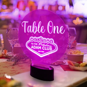 May include: A pink acrylic table number sign with the text "Table One" and a white neon sign that says "Welcome to the Fabulous Adam Club New York".