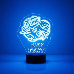 Pu&ograve; includere: Una luce notturna stampata in 3D blu e bianca con la mascotte dei Tar Heels della Carolina del Nord, un ariete, con il testo "UNC" e "ANY TEXT" sotto la mascotte.