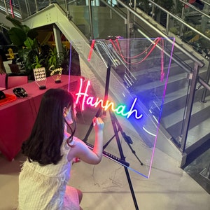 Op de afbeelding: Een jong persoon schrijft op een helder acryl bord met een regenboog neon-bord dat "Hannah" zegt.