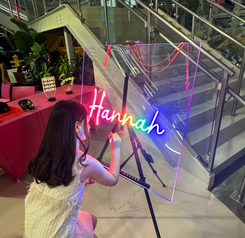 Op de afbeelding: Een jong persoon schrijft op een helder acryl bord met een regenboog neon-bord dat "Hannah" zegt.