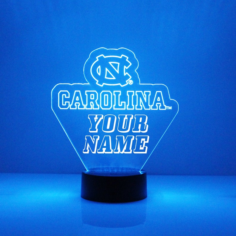 Pu&ograve; includere: Una luce notturna in acrilico bianco con un logo blu UNC Tar Heels e il testo "CAROLINA" e "YOUR NAME". La luce &egrave; su una base nera.