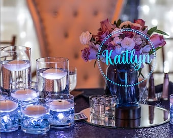 May include: A clear acrylic sign with a blue light and the name "Kaitlyn" in a white script font. The sign is on a table with a vase of pink and white flowers and several clear glass cylinders with floating candles.
