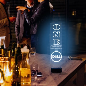 May include: A clear acrylic sign with the word "ONE" in white outlined letters. The sign also includes the text "CORPORATE CHRISTMAS PARTY 2020" and the Dell logo. The sign is lit from behind with a blue light.