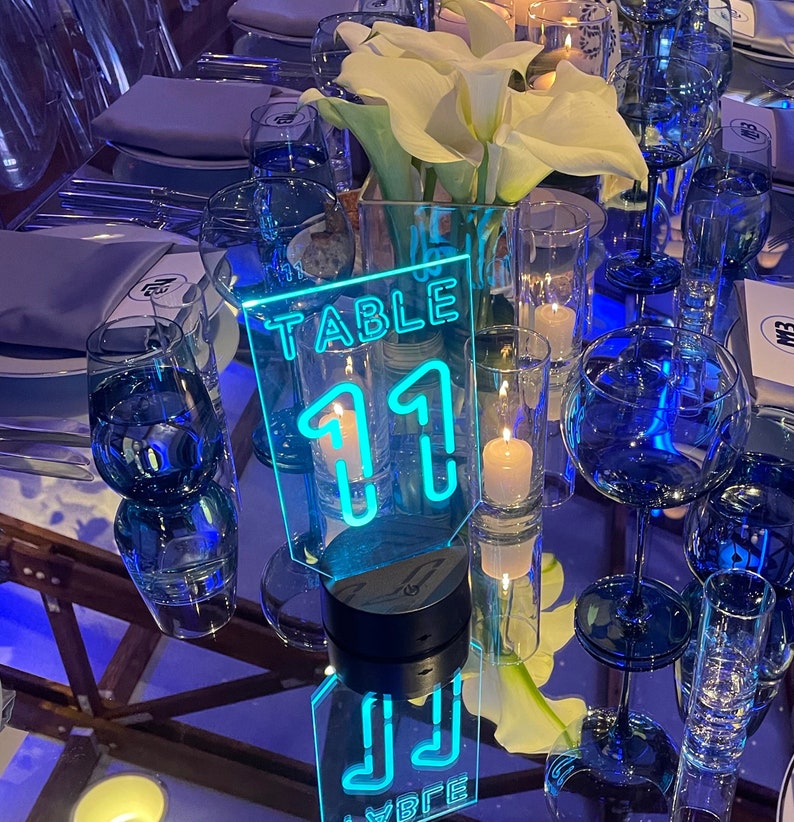 May include: A neon blue acrylic table number sign with the text "TABLE 11" in a modern font. The sign is on a black base and is illuminated from within.