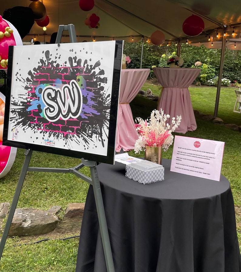 May include: A black easel with a white sign featuring a graffiti-style design with the letters "SW" in pink and blue. The sign is set up on a black table cloth with a small white box and a pink sign with the text "STOP" in white.