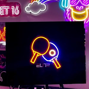 Op de afbeelding: Neon-bord met twee gele en rode pingpong-batjes, een blauw batje en het woord "ELITE" in witte letters op een zwarte achtergrond.