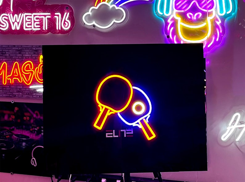 Op de afbeelding: Neon-bord met twee gele en rode pingpong-batjes, een blauw batje en het woord "ELITE" in witte letters op een zwarte achtergrond.