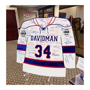 May include: A white, red, and blue acrylic hockey jersey sign featuring the name DAVIDMAN and the number 34 in dark blue. The surface is covered in handwritten signatures and messages, with two circular placeholders labeled YOUR LOGO HERE.