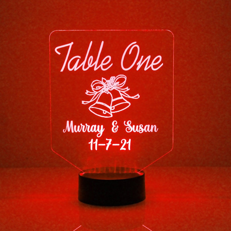 May include: A white acrylic table number sign with the text "Table One" and a wedding bell design. The sign also includes the names "Murray & Susan" and the date "11-7-21". The sign is illuminated with red LED lights.